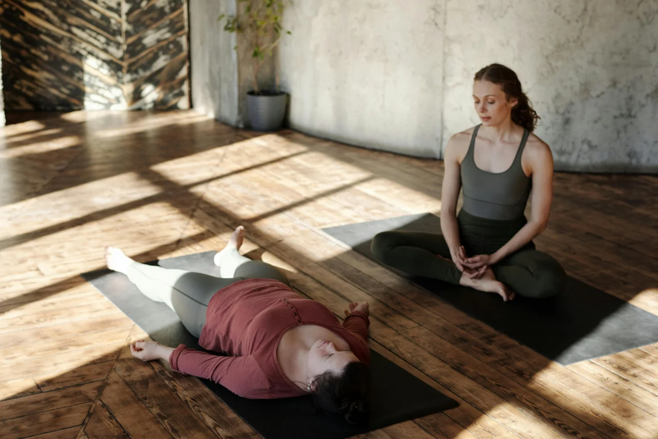 Meditation Rückenschmerzen 1 Patientin mit Rückenschmerzen liegt auf einer Matte und meditiert angeleitet von einer Frau neben ihr im Schneidersitz